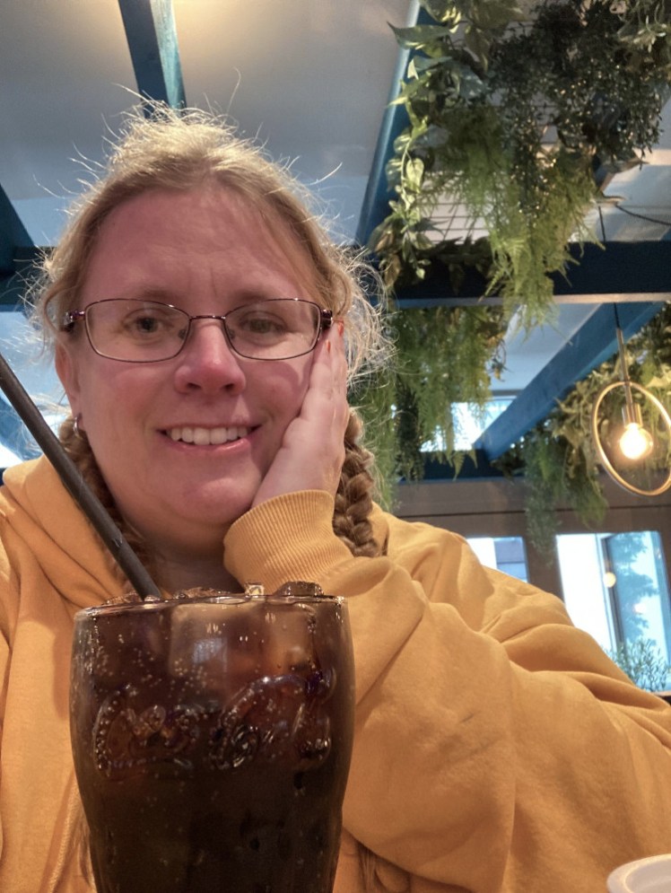 Me sitting in a pub with greenery hanging from the ceiling, wearing my yellow hoodie, with a pint of Coca-Cola in front of me.