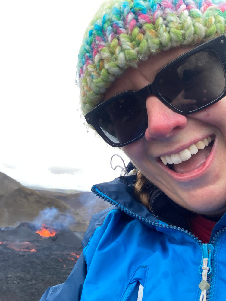 A very happy close-up selfie in raincoat, sunglasses and hat, lots of teeth visible. Over my shoulder you can see the volcano.