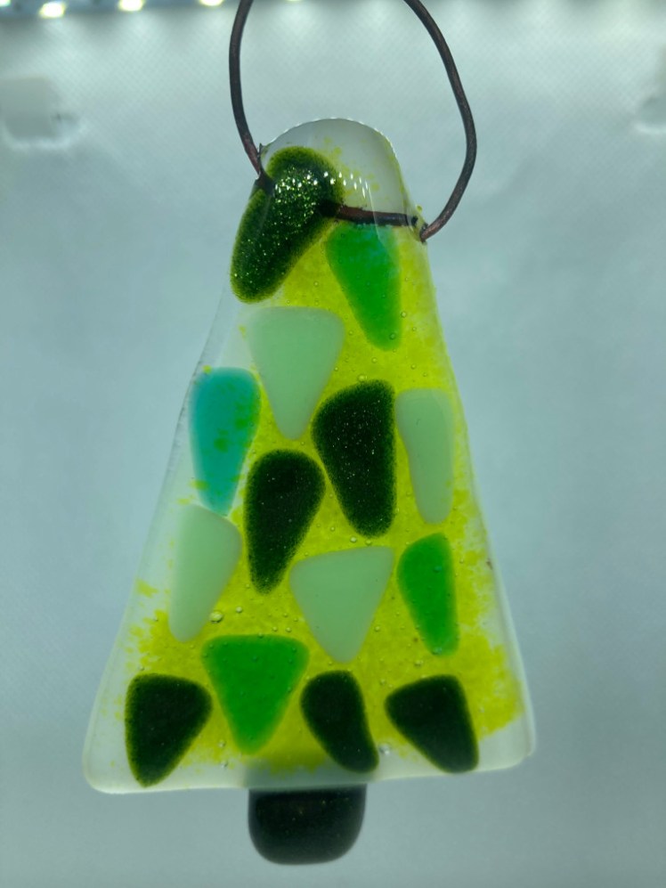 A fused glass Christmas tree about three inches tall with a loop of copper wire through the top for hanging. It's a base of a white glass triangle layered with green glass powder, small triangles in various shades of green and topped off with a clear glass layer. The whole thing has been melted together to become a thick solid hanging tree