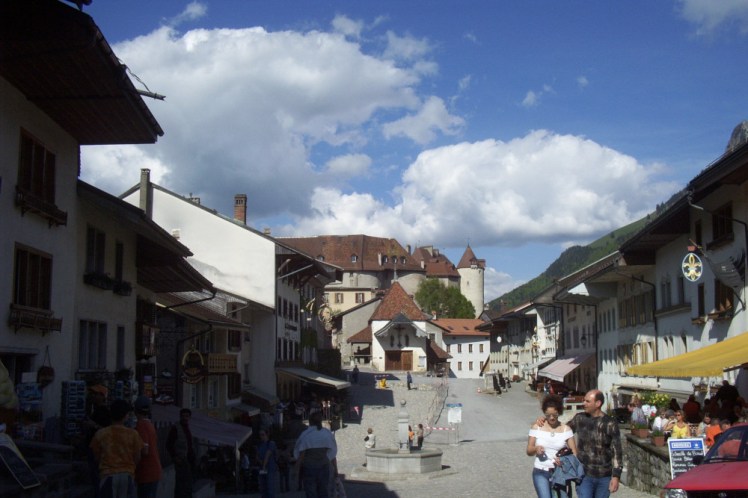 The centre of Gruyeres, looking across a cobbled square towards a castle. There are medieval-style chalet-like buildings down both sides of the square.