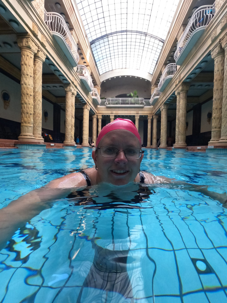 Me swimming in a beautiful pool surrounded by yellow stone columns and a glass roof while wearing a bright pink swimming cap.