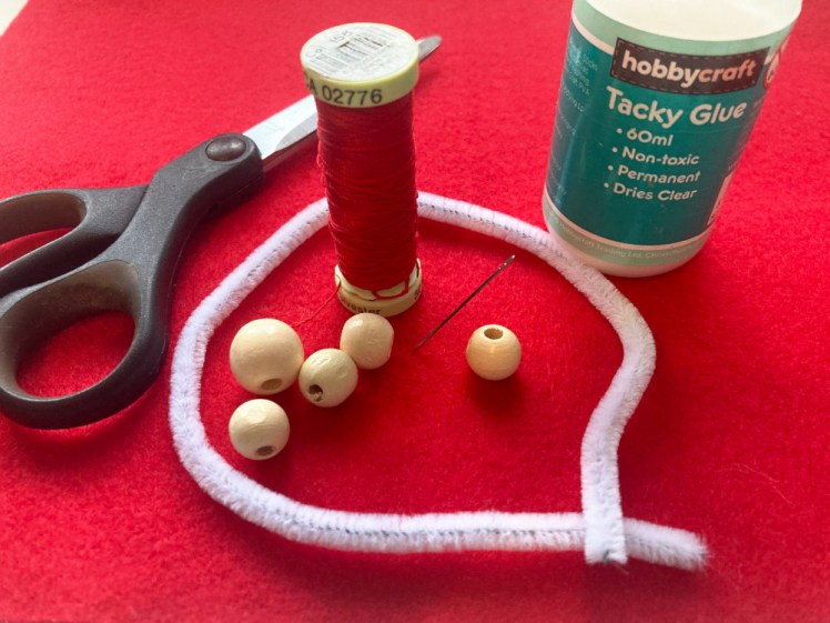 A pile of thread, scissors, needle, wooden beads, pipecleaner and pot of tacky glue lying on a sheet of red felt.