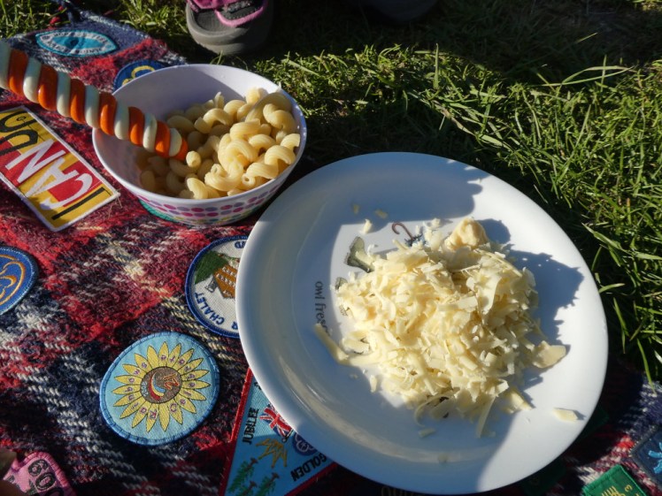 A bowl of spiral pasta and a plate of grated cheese, in the low evening sun, sitting on my camp blanket amid by 2006-2008-ish badges. In the pasta bowl is a fork with its handle wrapped in orange and cream polymer clay. The cream glows in the dark.