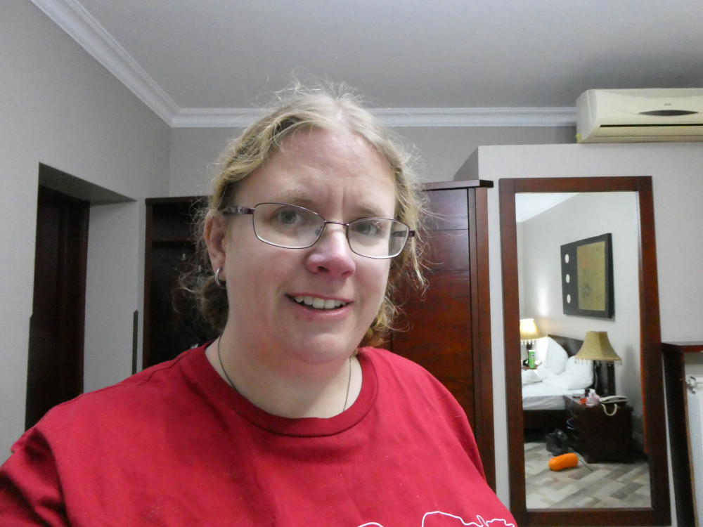 A selfie accidenally taken in the room when I meant to do a video. You can see half my stuff abandoned on or next to the bed. My hair is tied back rather than in plaits for once and I'm wearing a red t-shirt.