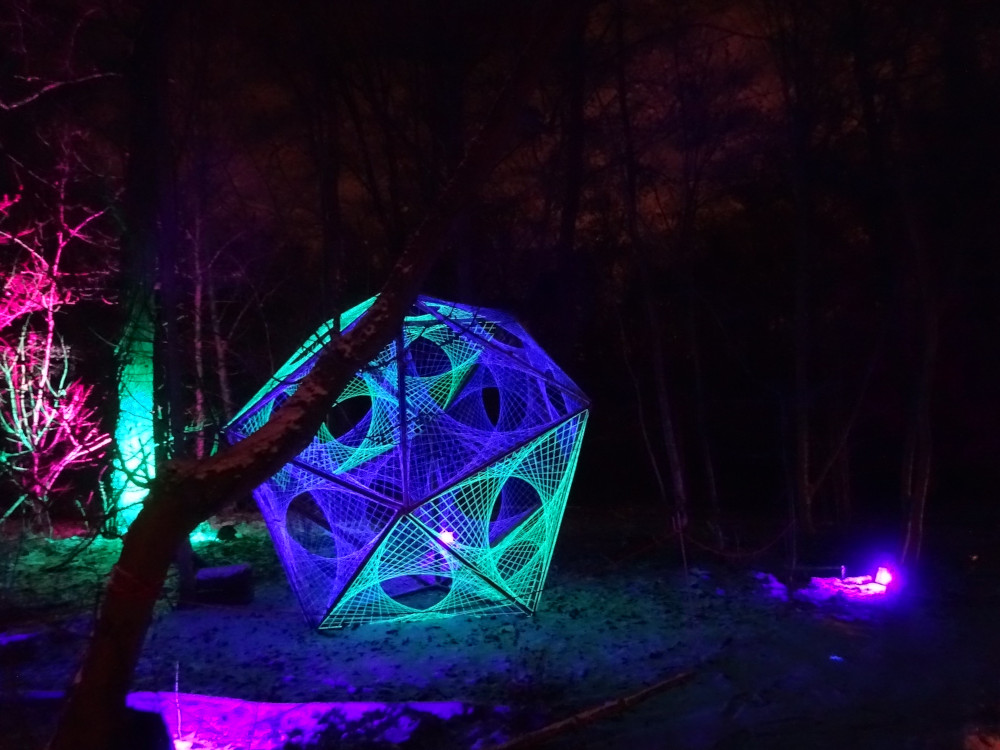 An irregular 3D shape, its faces made up of strings wrapped around to leave holes in the middle, more or less circular. It's lit by purple lights but the shape itself is green and blue and the green in particular looks like the strings themselves are glowing.