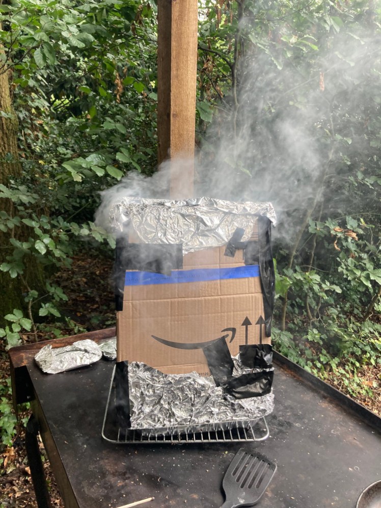 A cardboard box, lined with foil, sitting on top of a disposable barbecue with smoke billowing from the top.