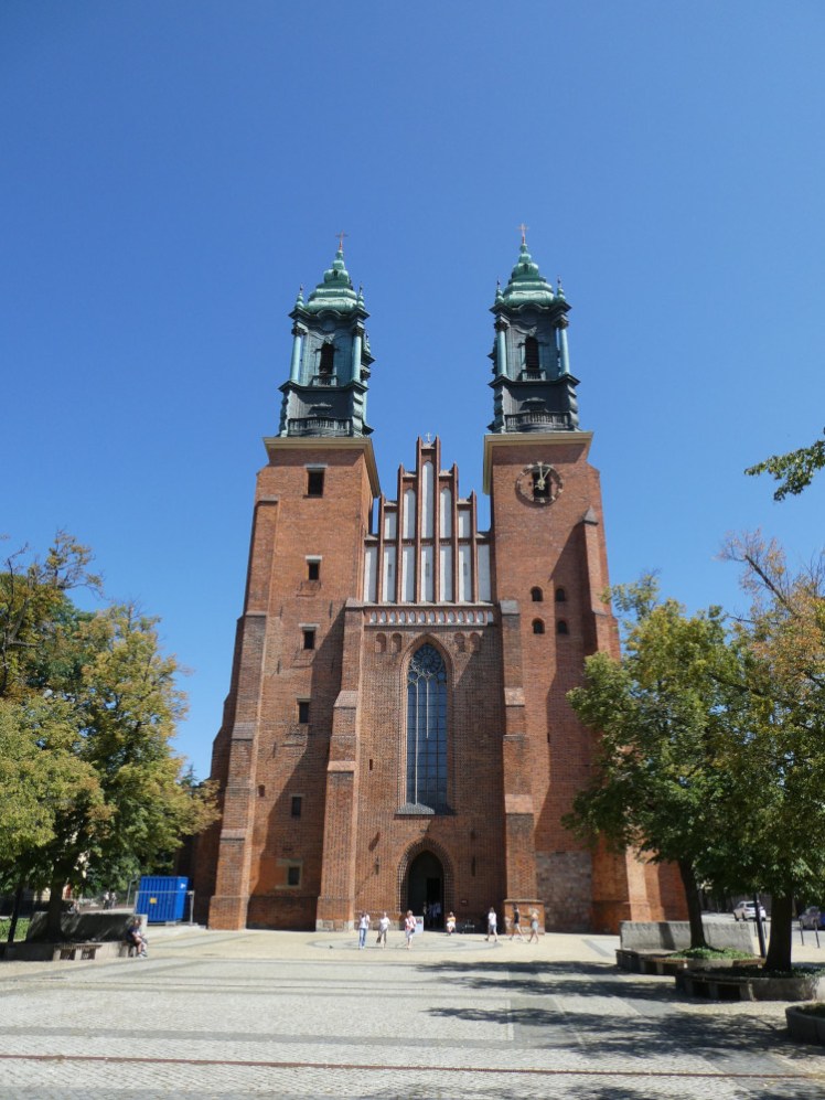 The exterior of Poznan Cathedral, a brick church with tall thin windows in the front, a tower on each side and a red and white ornate thing over the middle.