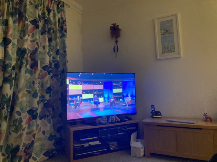 A photo of the TV glowing very blue in the corner of a dimly lit room. On the screen are the four House of Games contestants sitting in their chairs.