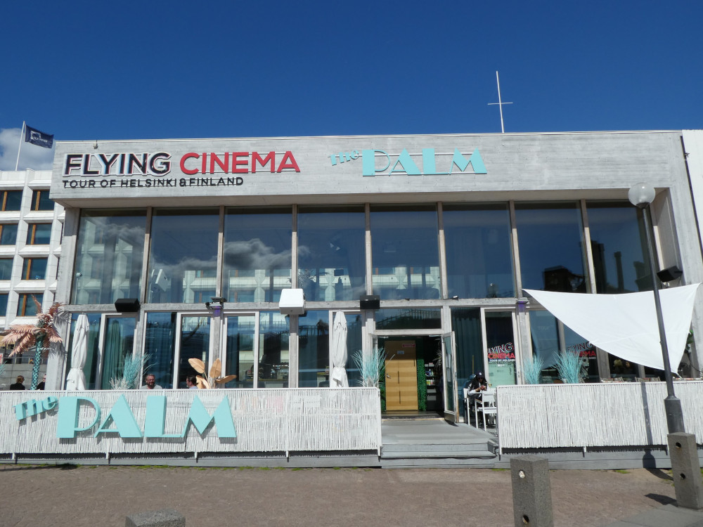 A grey concrete building that's actually mostly large glass windows. Along the top, it has the logo of Flying Cinema and also of The Palm in pale blue Art Deco letters, referring to the bar that occupies the other half of the square building.
