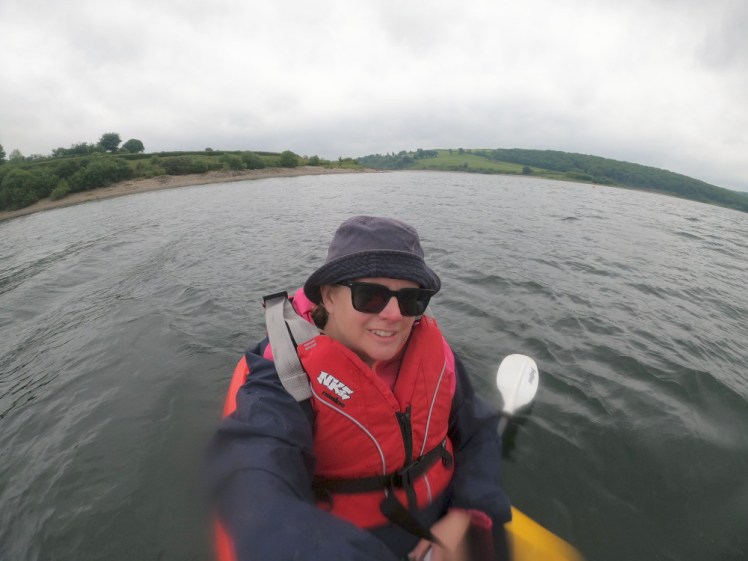 Me, wearing a red buoyancy aid and a blue bucket hat (because I didn't brush my hair in the morning and all the fluff was getting in my face) sitting in a kayak out in the lake. Behind me, green hills curve down to the lake, bent by the GoPro's lens.
