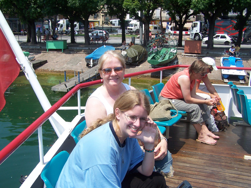 Renee, in a white vest top, and me, in a big pale blue t-shirt, on the back of the boat. We both look like this is the first time we've been out in the sun in ten years. Behind the boat, there are smaller boats pulled up onto the concrete edge of the lake, mostly with their covers on and behind a row of trees, you can make out the town.