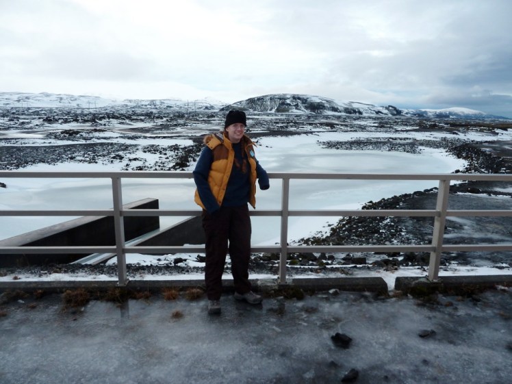 Me beside the frozen reservoir, leaning tentatively on the metal railings. I'm wearing a black fleece hat, blue fleece jumper and black hiking trousers with boots. Also an orangey-brown padded vest with a fur hood. It wouldn't do up even at the time. Behind me is a scene of black mountains covered reasonably thickly in snow and to my far right, you can just see the volcano Hekla.