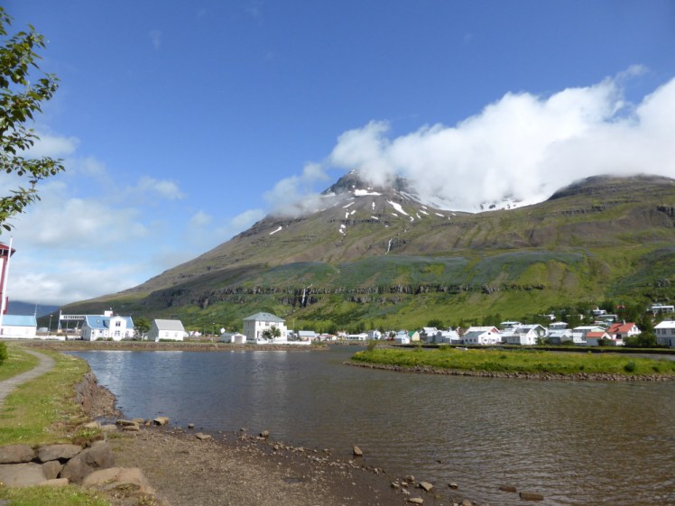 Seyðisfjörður as seen from beside a shallow river. The town is an assortment of houses, many of them white with red or blue roofs. A steep mountain looms over the town.
