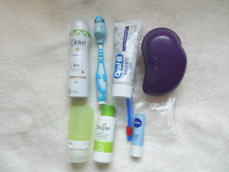 My washing kit. A small spray deodorant, a child's toothbrush in the shape of a shark, a tube of toothpaste, a purple Tangle Teezer hairbrush, a green silicon tube, a tiny tube of Simple moisturiser and a blue lipbalm, all laid out on top of a plastic sandwich bag. 