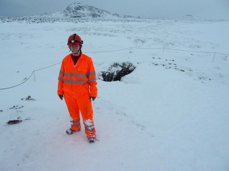 Me, post-caving, standing in the snow outside a hole in the snow which is the cave entrance. I'm wearing a red helmet with a torch on it and a neon orange boilersuit with reflective stripes across it.