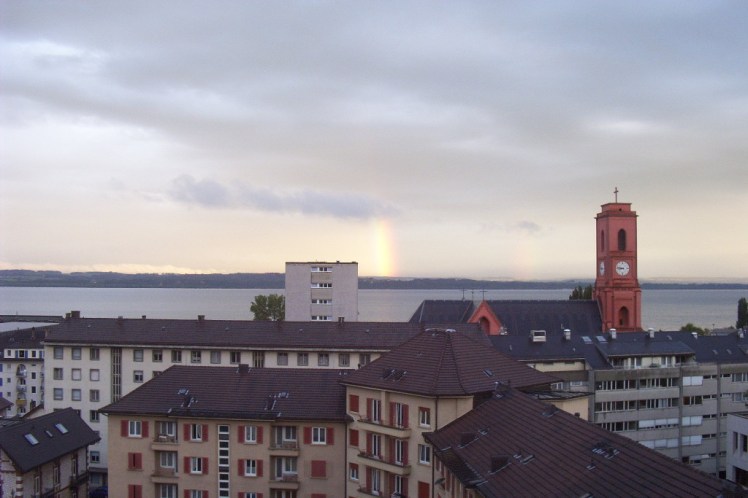 The view from the window of my room in Neuchatel. In the distance there are mountains, beyond a lake. In the foreground are apartment buildings and in between is a red church with a tower.