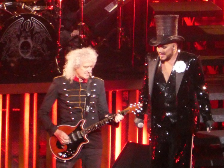 Brian May and Adam Lambert. May is wearing the military jacket and playing the guitar. Lambert is wearing a black sequinned suit and a top hat.