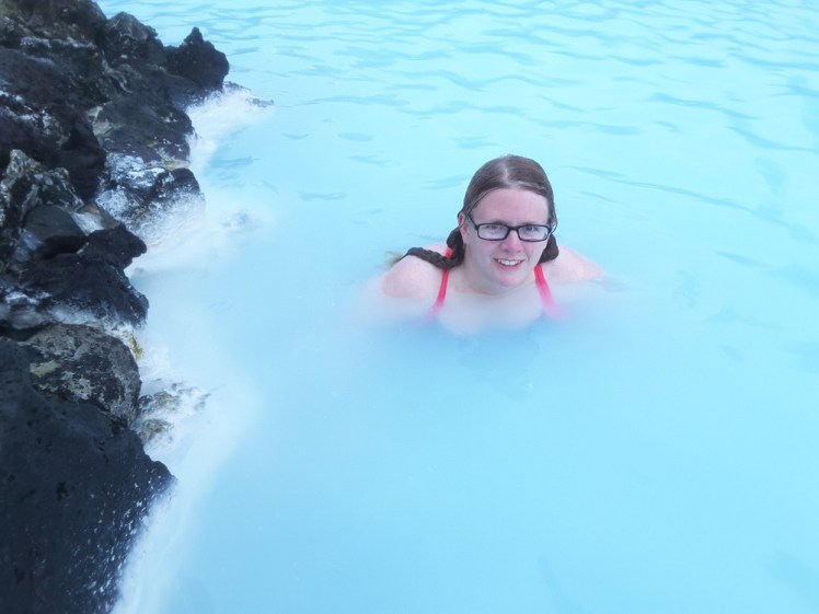 Me, in the Blue Lagoon, only my head and shoulders visible in the opaque bright blue water. The photographer has positioned me so that the silica-streaked black lava rocks that form the side are framing the left side of the picture.