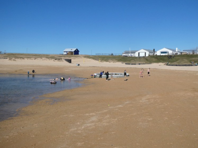 People on the beach around the hotpot, clearly showing where the warm patch is in the water. Some are in swimwear, some are in winterwear and some are kids playing on giant tyres in the water.