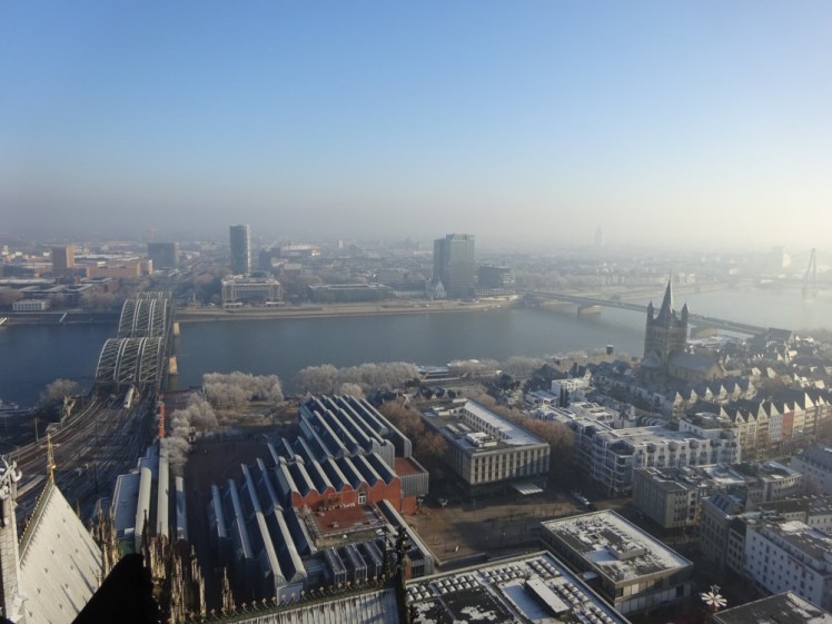 A view over Cologne from the cathedral tower. To the right is a three-lane arched railway bridge crossing a wide blue river. In the foreground is another church with a lower Gothic tower and you can make out Hanseatic-style buildings just below that. There's definitely a thick haze coming in from the west.