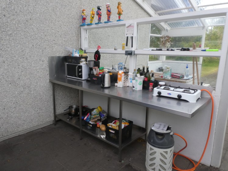 Hveragerði campsite kitchen - a gas stove tethered to a huge canister, a shelves of food, a microwave, extension leads for charging stuff and behind the divider, benches visible, all under a clear plastic roof. The wall of the kitchen is clearly the outside wall of the solid building next door.