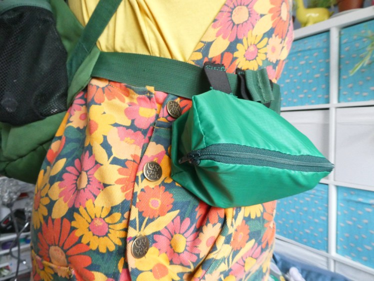 My mid-section turned sideways, wearing a yellow top and orange flowered dungarees. I'm wearing a green backpack. On the waist belt is a dark green rectangular pouch which is hanging awkwardly from two elastic loops.