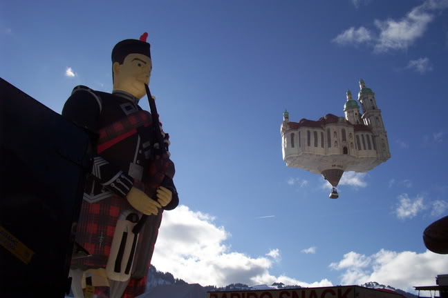 A Scottish piper-shaped balloon about to take off, while a cathedral-shaped balloon floats off to his left.