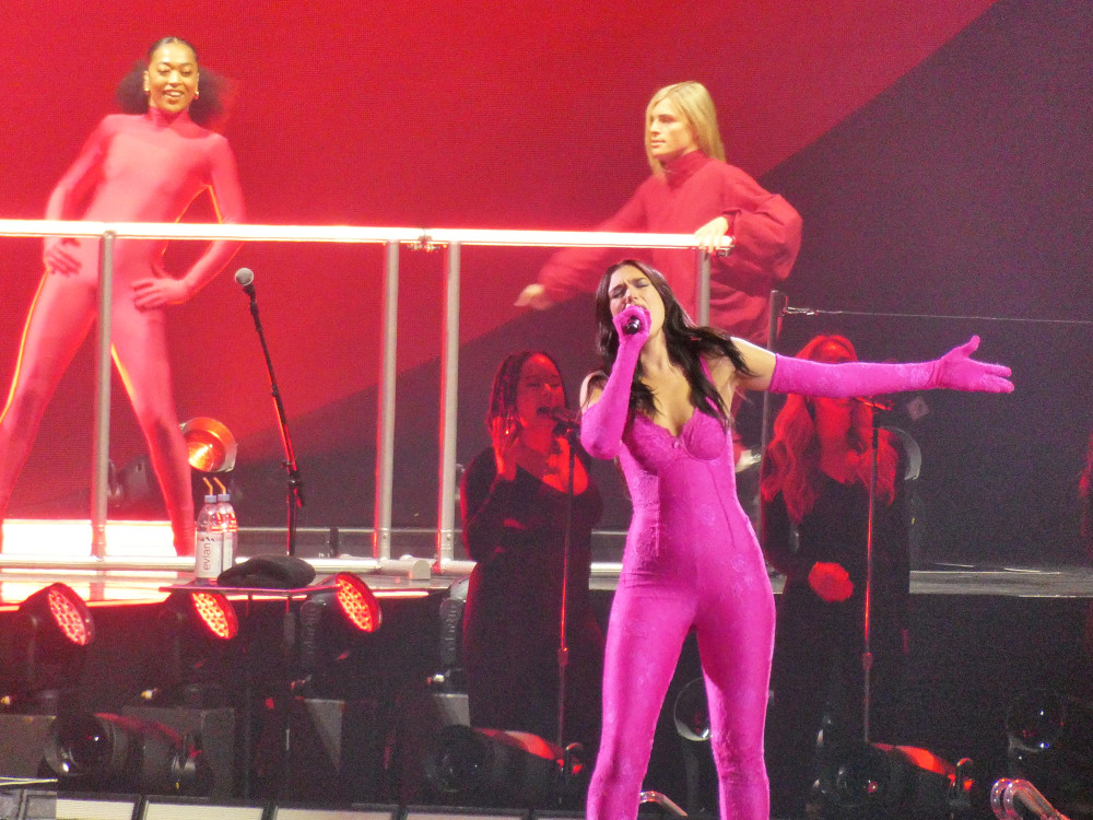 Dua Lipa on stage in Liverpool. I'm actually at the far end of the arena but this is a good close-up photo with a good zoom. Dua is wearing a neon pink catsuit with matching opera-length gloves. Even at this distance, you can see how the suit is actually lacy.