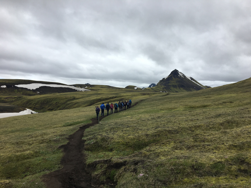 A dirt track crosses a dull green landscape. On the trail, eight or nine people are walking in a a line towards a pointy mountain and a heavy cloud in the distance.