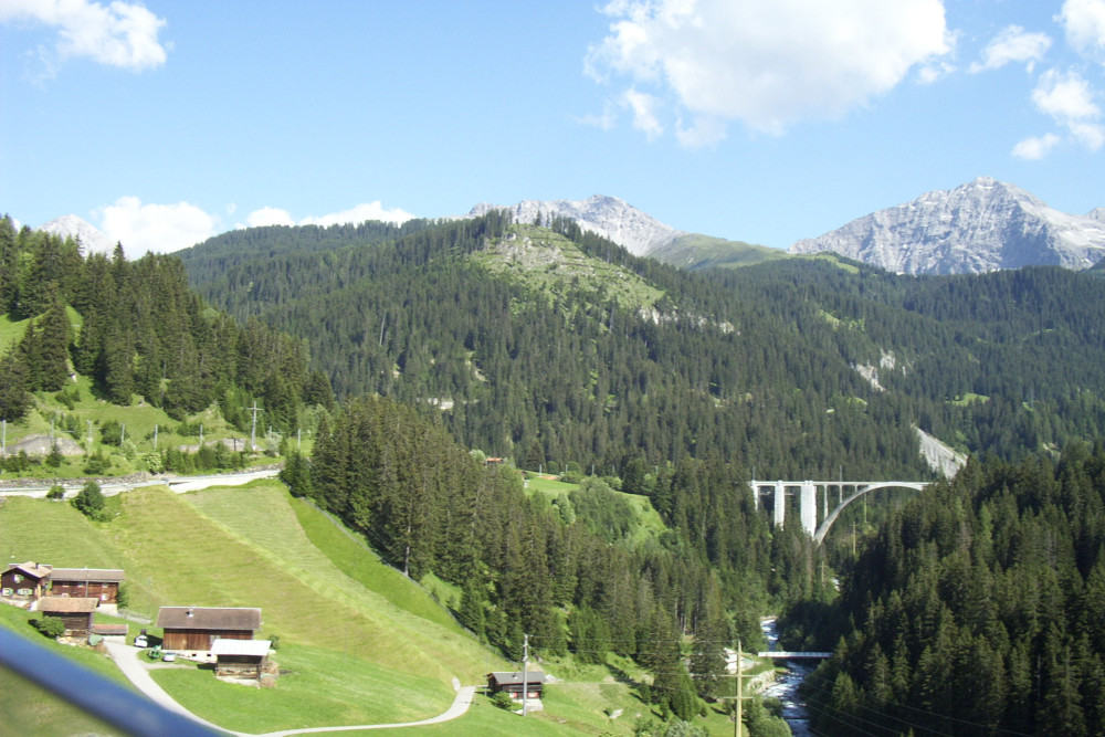 A beautiful Swiss view taken from a moving train. Bare rocky pyramid-shaped mountains poke out of lower pine-covered mountains. In the foreground is a steep-sided valley crossed by an interesting bridge among the trees.