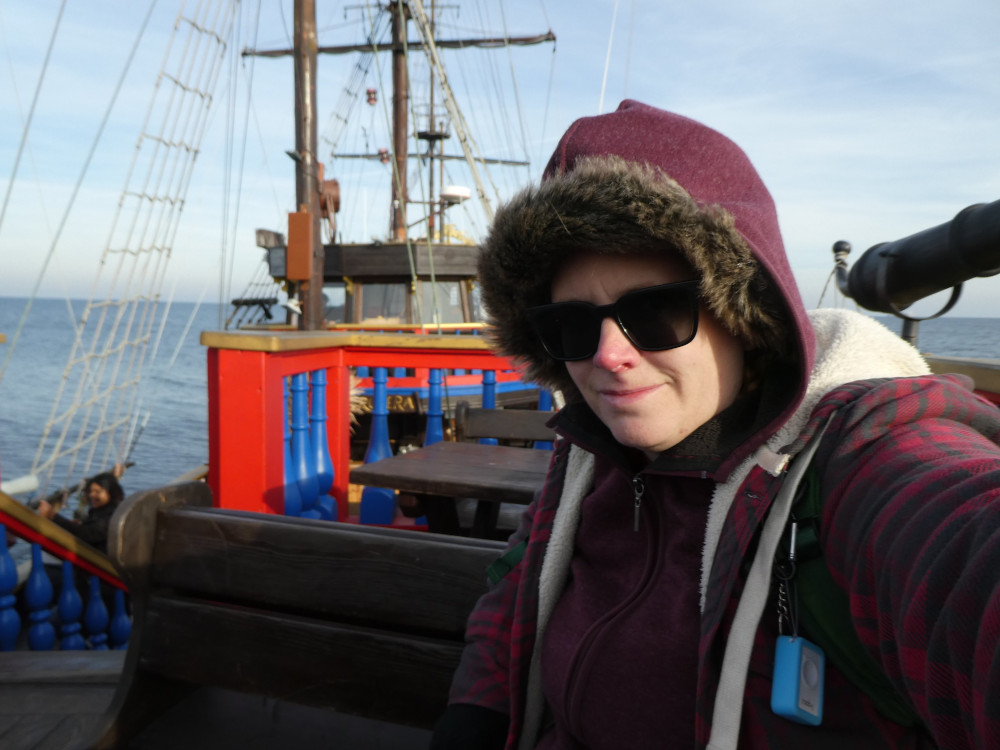 Selfie on a bright-painted fake pirate ship in north Poland in November, wearing a furry hat, a hoodie with the hood up and a slightly thicker button-up fleece shacket/hoodie. I'm as cold as I look.