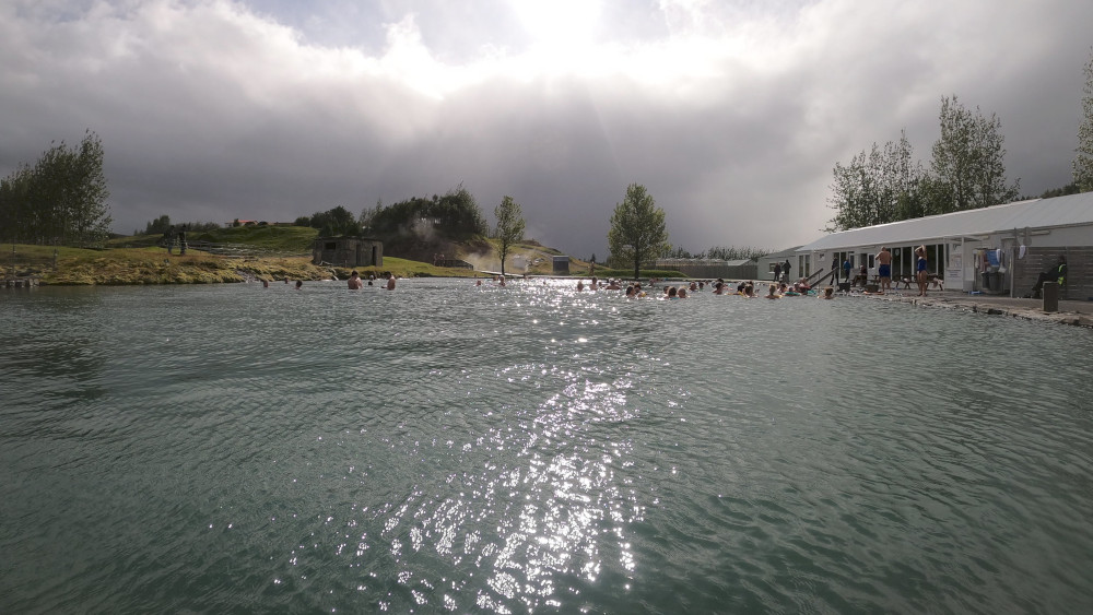 Secret Lagoon. A pool surrounded by hillocks and trees and its own geysers, all a lot more rustic-looking than the others. There is heavy cloud but a hint of sun is reflecting on the rippled water.