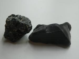 Two pieces of black rock, both raw obsidian. It's not as shiny as the processed version. One is a pitted cube of rock and one's a smoother sharper piece that looks a bit like it's been broken rather than worn.