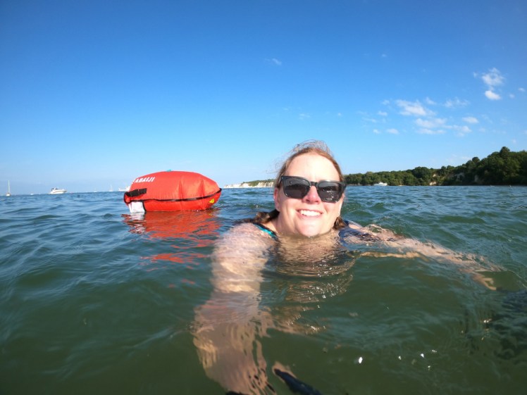 Sea swimming with the Handler mount. I'm swimming in a greenish-blue sea reflecting the bright blue sky overhead, with an orange tow float behind me and my right arm held out to take the photos.