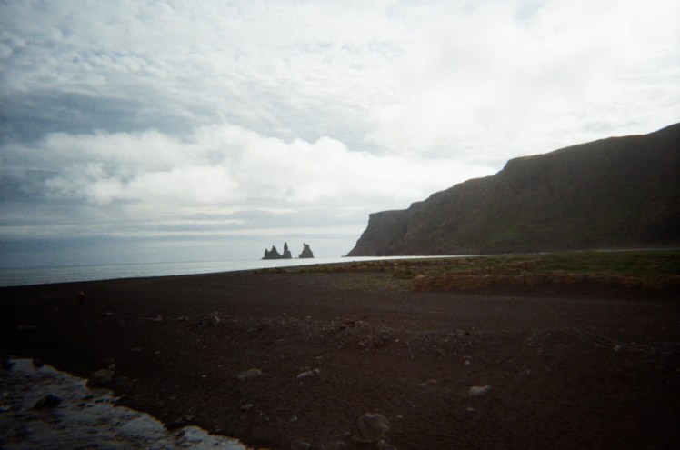 A large black headland behind a black beach. Just out at sea are several very pointy pinnacles.