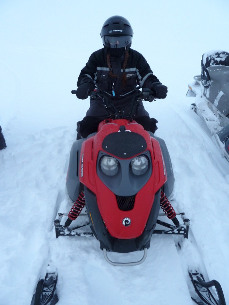 Me on a snowmobile. It's red and quite small. I'm wearing an all-in-one black suit and a helmet with a black visor.