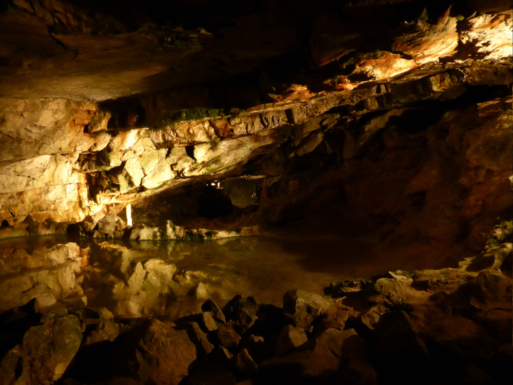 A cave lit in yellow by hidden electric lights. The roof is low, sloped and blocky and below is a lake, nearly reflecting the ceiling.
