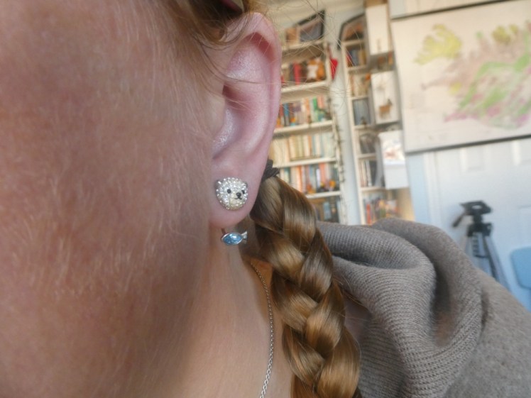 A close-up of my left ear and the fur on my cheek that's not as visible from further away. The earring is a silver crystal polar bear head with a blue crystal fish hanging from a hook attached to the back. The earring you can't see is a blue fish with a polar bear head dangling from an inch or so of chain.