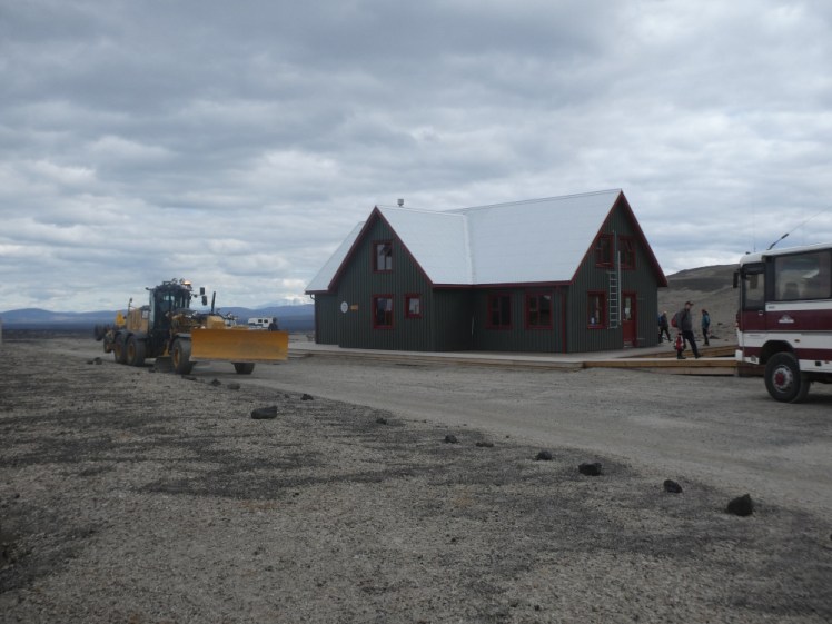 The cafe hut at Dreki, a dark-green building with a pointed roof. In the foreground is an enormous road-scraping machine and our coach is just peeking into the corner.