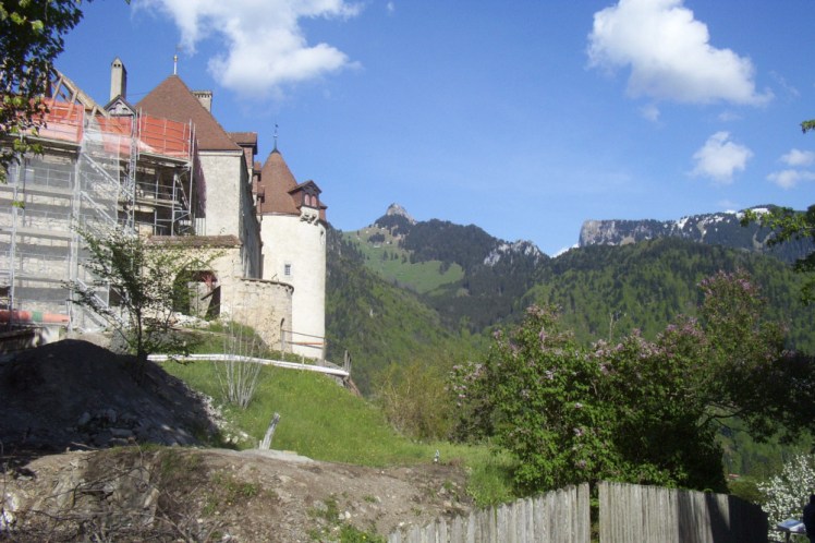 Gruyeres Castle, partly covered in scaffolding. Behind it you can see craggy mountains with green rolling foothills.
