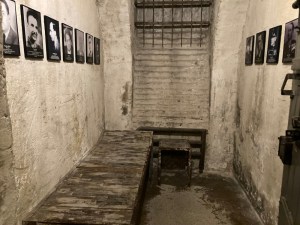 A basement cell. It's whitewashed but patchily, there's a rickety mouldy-looking wooden bed on one side and on the walls are pictures of people who would have been held in these cells.
