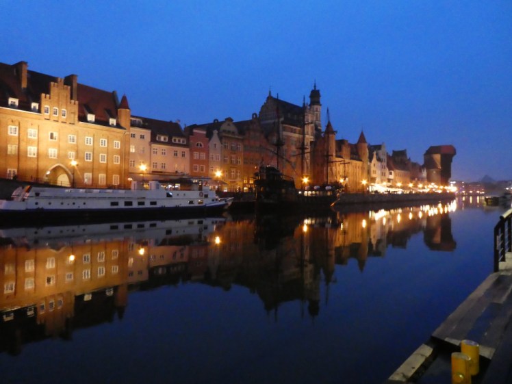 Brick warehouses along the edge of the river, lit up in yellow lights, reflect in the deep blue water reflecting the deep blue evening sky.