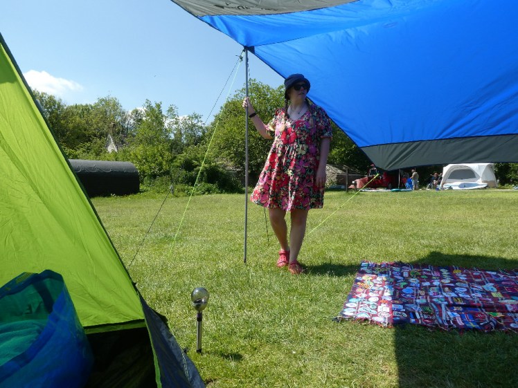 Me, in a short flowery dress, standing outside my green tent, under a blue tarp on a sunny day. My camp blanket is on the floor next to me.