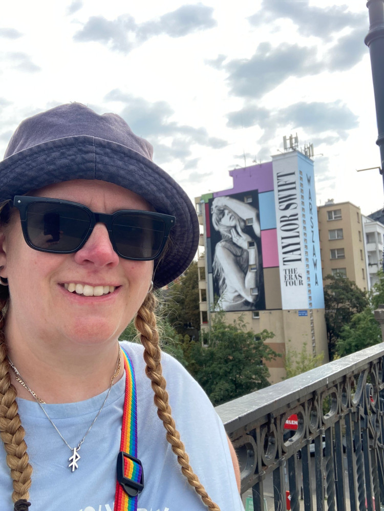 A selfie with the Eras mural, which is on the side of a small apartment building behind me. It features the words Taylor Swift: The Eras Tour vertically down the right hand side, with Warszawa just visible around the edge of the building. THe main part of the mural is a big painting of Taylor Swift in her Anthology pose and then there are squares painted in the Eras colours around her.