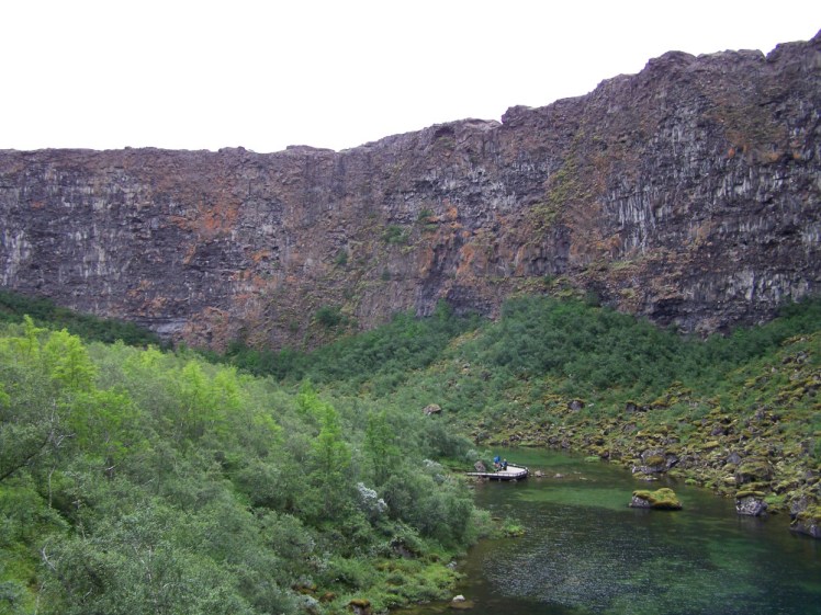 A huge basalt cliff with shrubs and a pond at the bottom. The cliffs don't look that high until you spot the tiny specks of people on a wooden platform sticking into the pond.