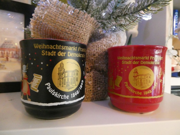Two ceramic mugs under a small Christmas tree. One is matt black, one shiny red but they both have a gold circle containing a picture of a church, a reindeer holding a scroll on the left and Santa waving a German flag on the right.