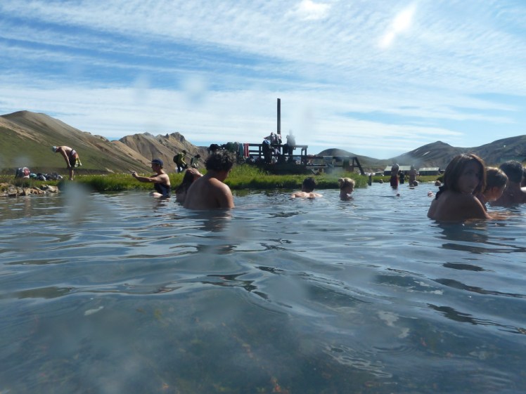 The Landmannalaugar hot pool. The sky is blue and the photo is taken from low down, in the water. People are sitting around in the water. Behind the pool is a wooden structure where you change and leave your clothes and behind that, you can see orange-streaked mountains.