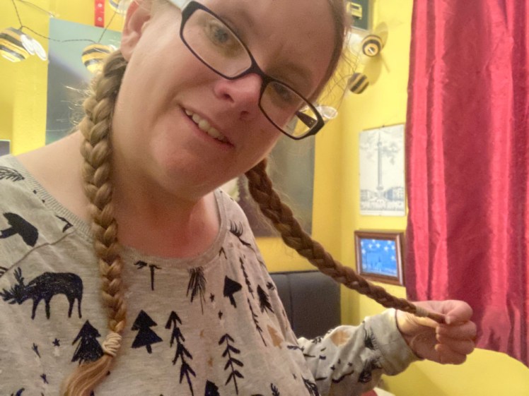 A selfie holding out one plait. I am wearing a grey pyjama top with pine trees and reindeer on it. Behind me are some very bright yellow walls and very red silky (polyester!) curtains.