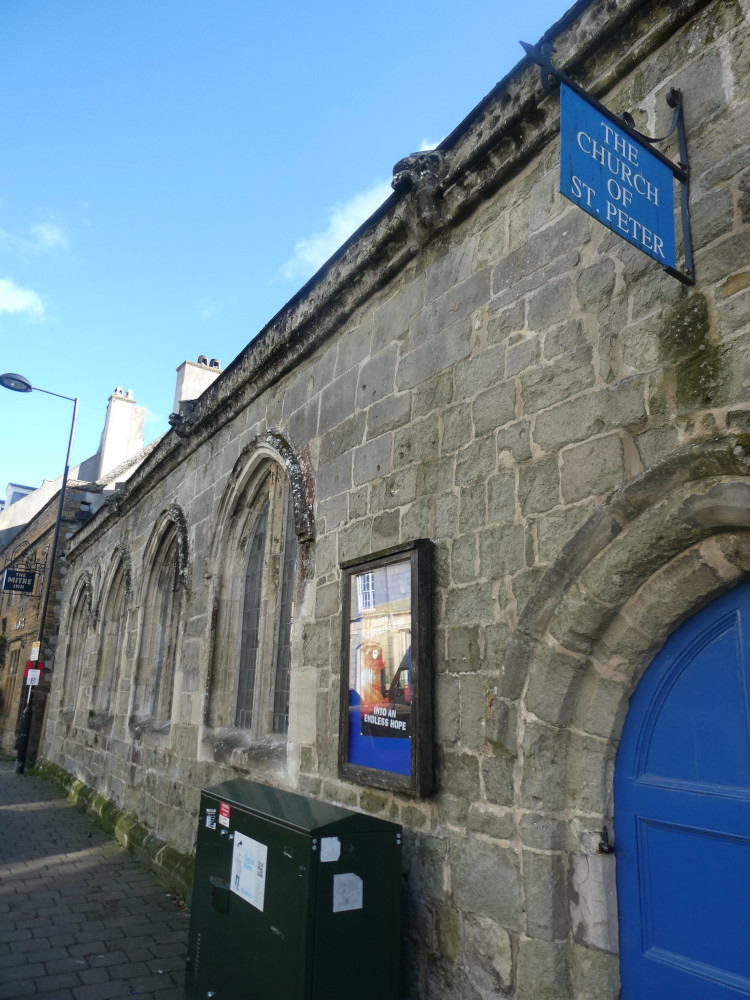 The exterior of St Peter's Church, an ancient stone wall with arched windows and door. From this angle, it's quite low but there's more above and behind me is the tower, which you also can't see.