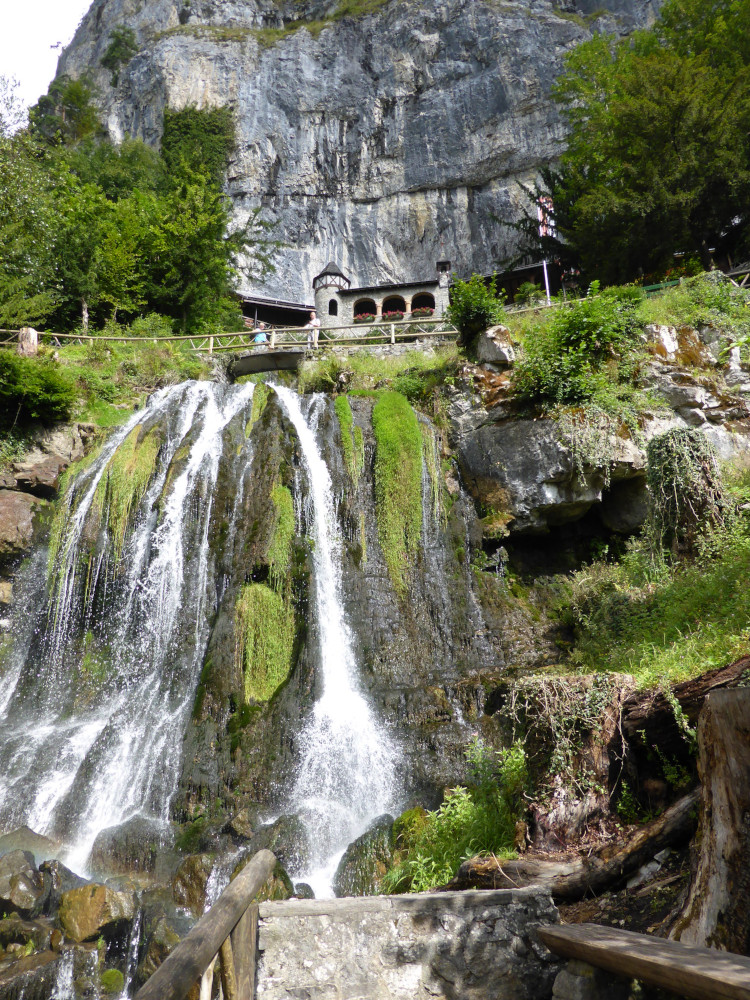 The entrance to St Beatus Caves from below. A waterfall flows over mossy rocks and there's a path with wooden handrails leading up to a stone frontage which has a squat tower and some arches.